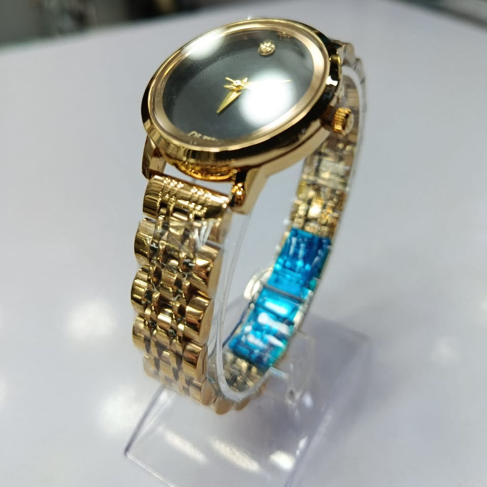 A gold-tone women's watch with a dark gray dial and a multi-material link bracelet featuring gold and blue textured segments, displayed on a clear stand.