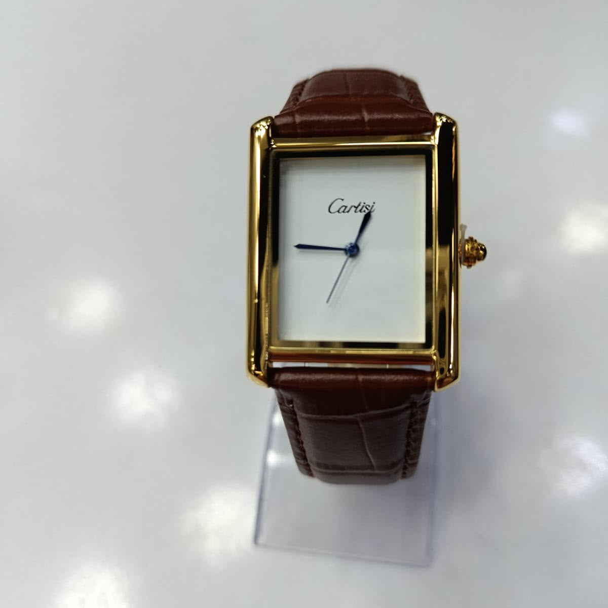 A Cartier Tank watch with a gold-tone rectangular case, white dial, and brown leather strap, displayed on a clear stand against a white background.