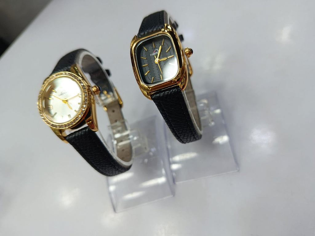 Two gold-tone wristwatches with black textured leather straps displayed on clear acrylic stands against a white background.