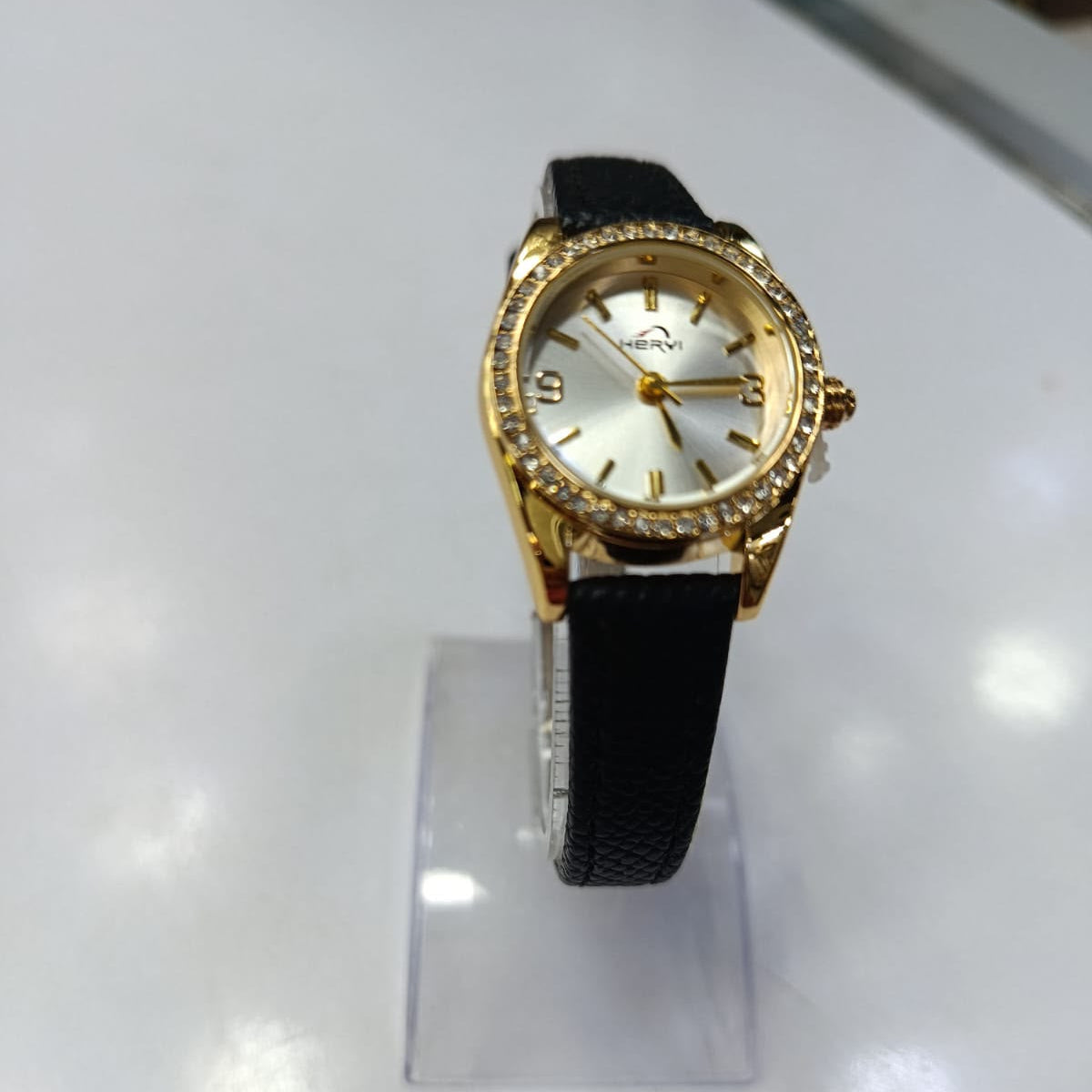 Omega women's watch with a gold-tone case, crystal-studded bezel, white dial, and black textured strap, displayed on a clear stand.