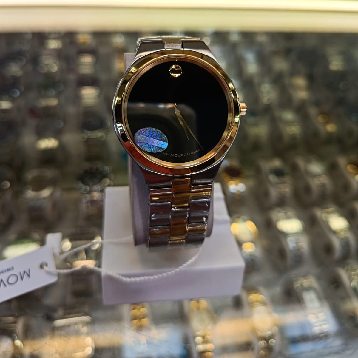 A Movado watch with a gold-tone metal case and bracelet, featuring a black dial with the signature dot-motor movement indicator, displayed on a white stand inside a watch store.