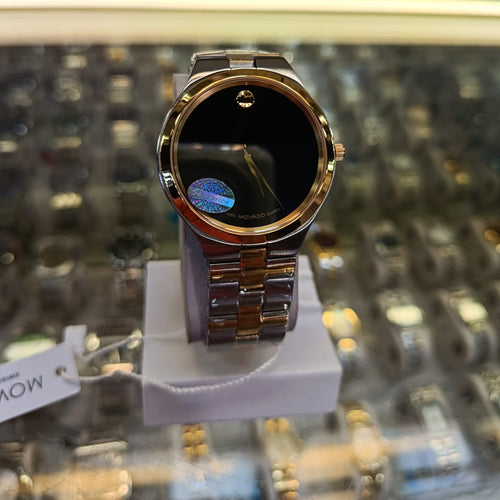 A Movado watch with a gold-tone metal case and bracelet, featuring a black dial with the signature dot-motor movement indicator, displayed on a white stand inside a watch store.