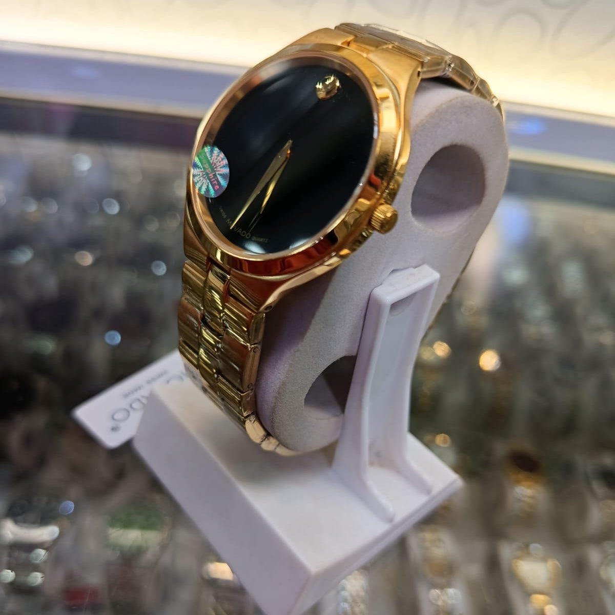 A gold-tone metal watch with a black dial and a small round iridescent crystal accent, displayed on a white stand inside a watch shop.
