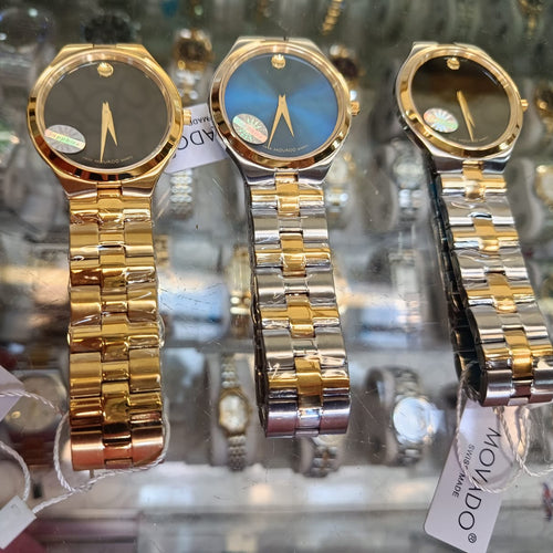 Three Movado wristwatches displayed side by side in a glass case, featuring gold and silver metal bracelets and round dials in black and blue.